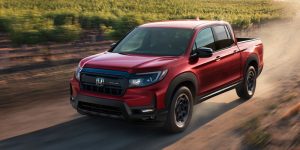 A red 2025 Honda Ridgeline driving on a scenic dirt road surrounded by vineyards.