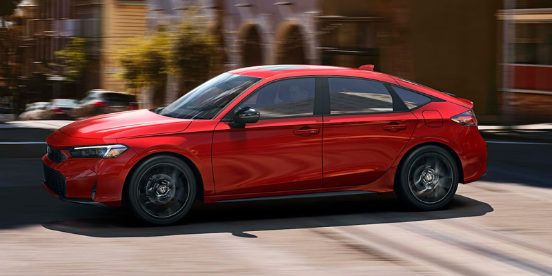 Side view of a red sedan driving through a city street with motion blur in the background, highlighting its sleek profile and sporty design.
