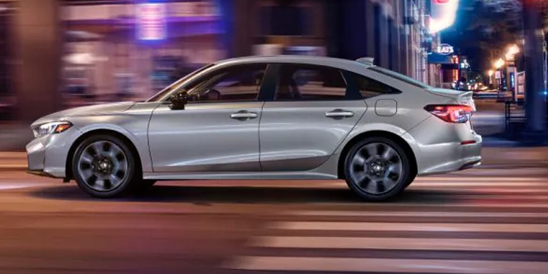 Side view of a silver sedan driving through a city street at night, with motion blur and illuminated urban lights in the background as it passes over a crosswalk.
