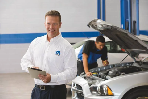 Service advisor holding a tablet in a dealership service center while a technician performs maintenance on a vehicle in the background.